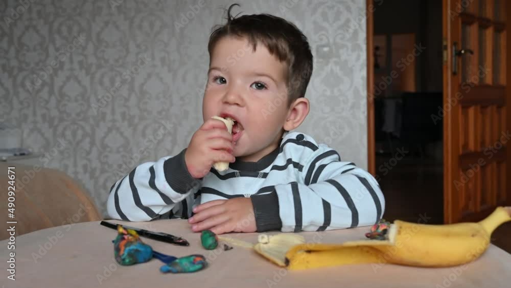 Funny caucasian toddler boy at the table eating a banana. Snack during the game Funny caucasian toddler boy at the table eating a banana. Snack during the game