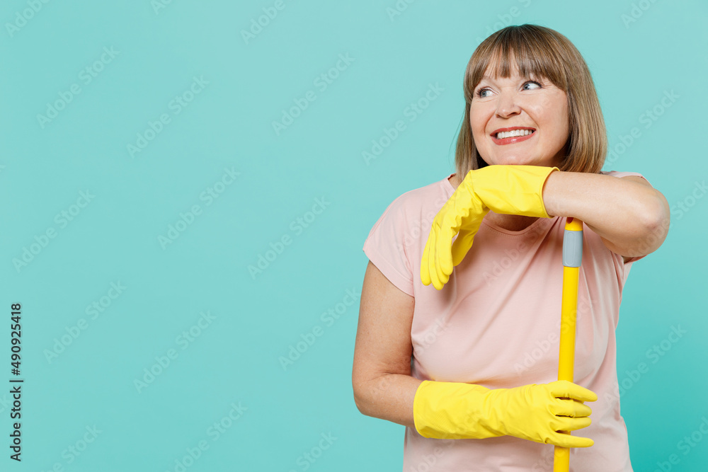 Elderly housewife woman 50s in pink t-shirt gloves do housework leaning on mop look aside on workspace mock up isolated on plain pastel light blue background Housekeeping cleaning tidying up concept. Elderly housewife woman 50s in pink t-shirt gloves do housework leaning on mop look aside on workspace mock up isolated on plain pastel light blue background Housekeeping cleaning tidying up concept.