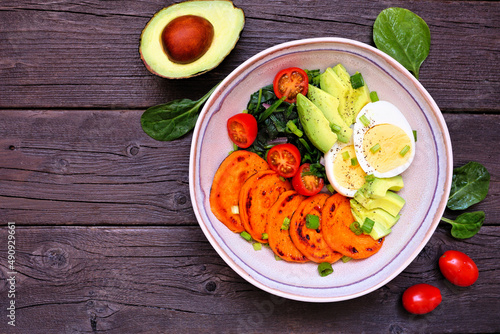Nutritious breakfast bowl with sweet potato, egg, avocado and spinach. Above view table scene on a dark wood background.