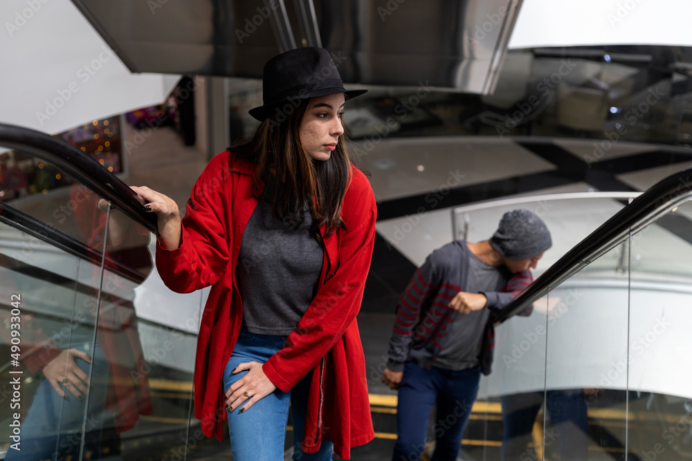 Two young Latin American brothers walk up escalators in a shopping mall. She wears a black hat and red sweater, he wears a hat and gray sweater. Walking and leisure concept.