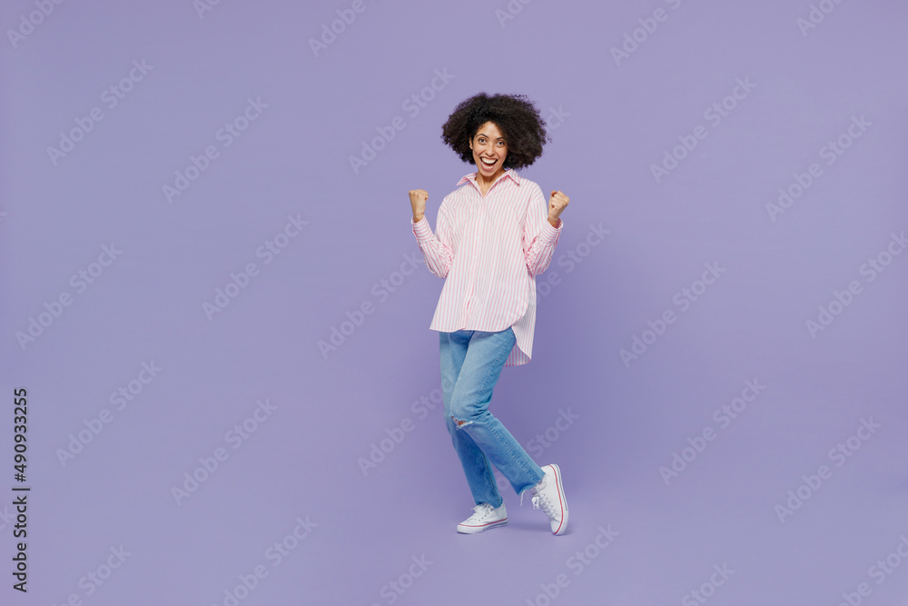 Full body young overjoyed fun woman of African American ethnicity 20s wear pink striped shirt doing winner gesture celebrate clenching fists say yesisolated on plain pastel light purple background Full body young overjoyed fun woman of African American ethnicity 20s wear pink striped shirt doing winner gesture celebrate clenching fists say yesisolated on plain pastel light purple background