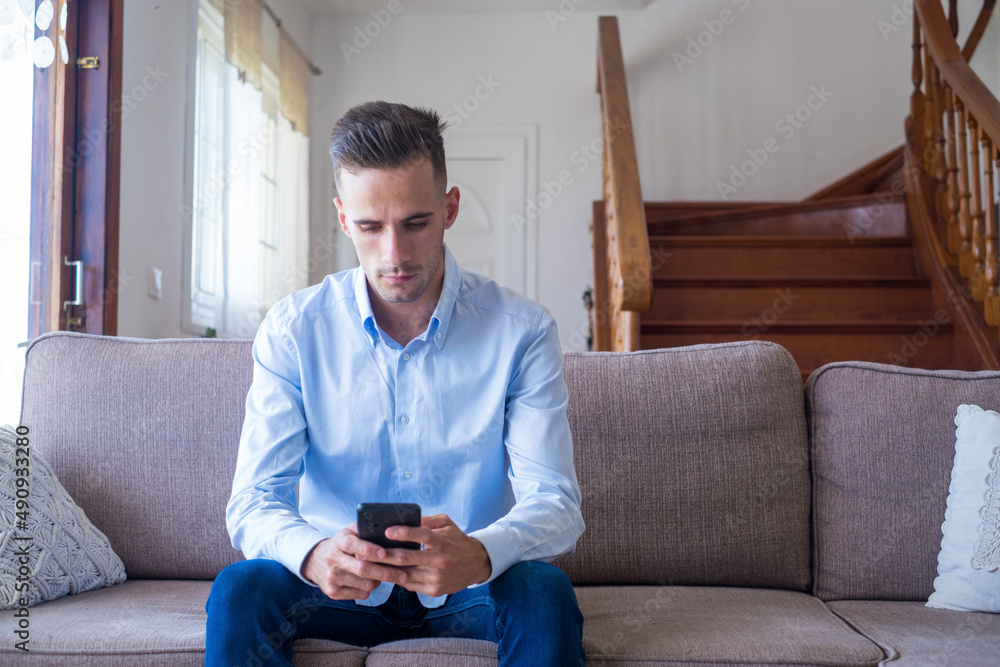 Serious young man sitting on sofa and text messaging using mobile phone at home. Handsome caucasian guy browsing social media content or chatting online on smartphone while relaxing on couch Serious young man sitting on sofa and text messaging using mobile phone at home. Handsome caucasian guy browsing social media content or chatting online on smartphone while relaxing on couch