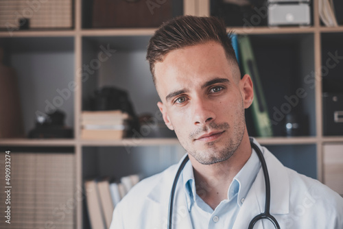 Portrait of handsome young male doctor in white lab coat with stethoscope looking at camera from medical office. Caucasian male confident healthcare worker in uniform at workplace
