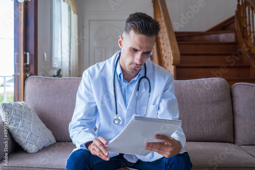 Portrait of handsome young male doctor in white lab coat with stethoscope looking at reports in medical office. Caucasian male healthcare worker in uniform analyzing written report or test results  photo