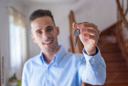 Portait of smiling young man showing or giving keys of his new house. Confident handsome male holding keys of the home or apartment in living room. Concept of buying or selling a property