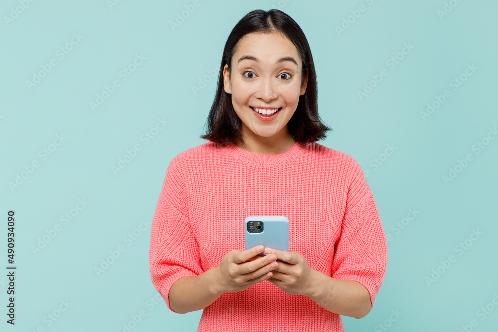 Young smiling cool happy woman of Asian ethnicity 20s wearing pink sweater hold in hand use mobile cell phone isolated on pastel plain light blue background studio portrait. People lifestyle concept. Young smiling cool happy woman of Asian ethnicity 20s wearing pink sweater hold in hand use mobile cell phone isolated on pastel plain light blue background studio portrait. People lifestyle concept.