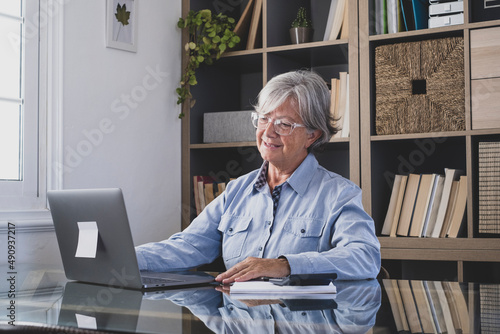 Senior businesswoman video chatting online using laptop with adhesive note at home office in front of book shelf. Elderly caucasian woman working online or watching media content at workplace