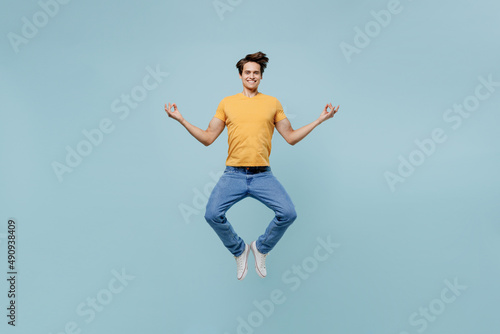 Full body spiritual young man wear yellow t-shirt jump high hold spreading hands in yoga om aum gesture relax meditate try to calm down isolated on plain pastel light blue background studio portrait