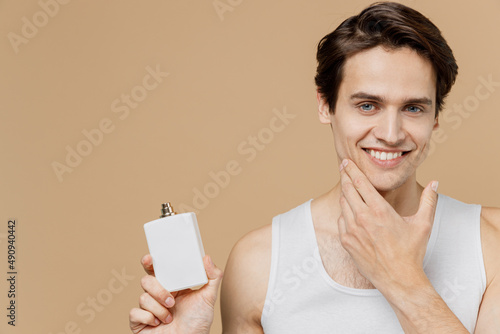 Attractive happy young man 20s perfect skin wear undershirt hold aftershave deodorant bottle touch chin isolated on pastel pastel beige background. Skin care healthcare cosmetic procedures concept photo