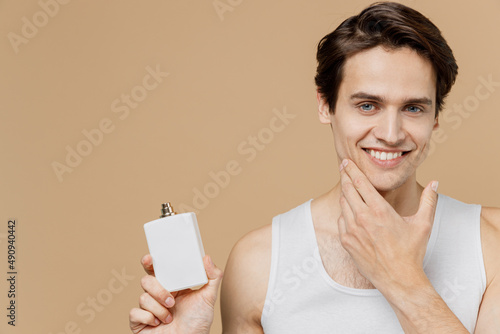 Tableau sur toile Attractive happy young man 20s perfect skin wear undershirt hold aftershave deodorant bottle touch chin isolated on pastel pastel beige background