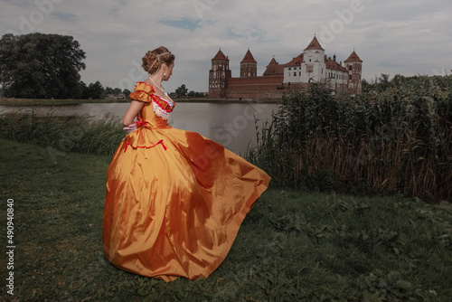 Fotografie A young woman in an old orange dress walks on the shore of the lake near the cas