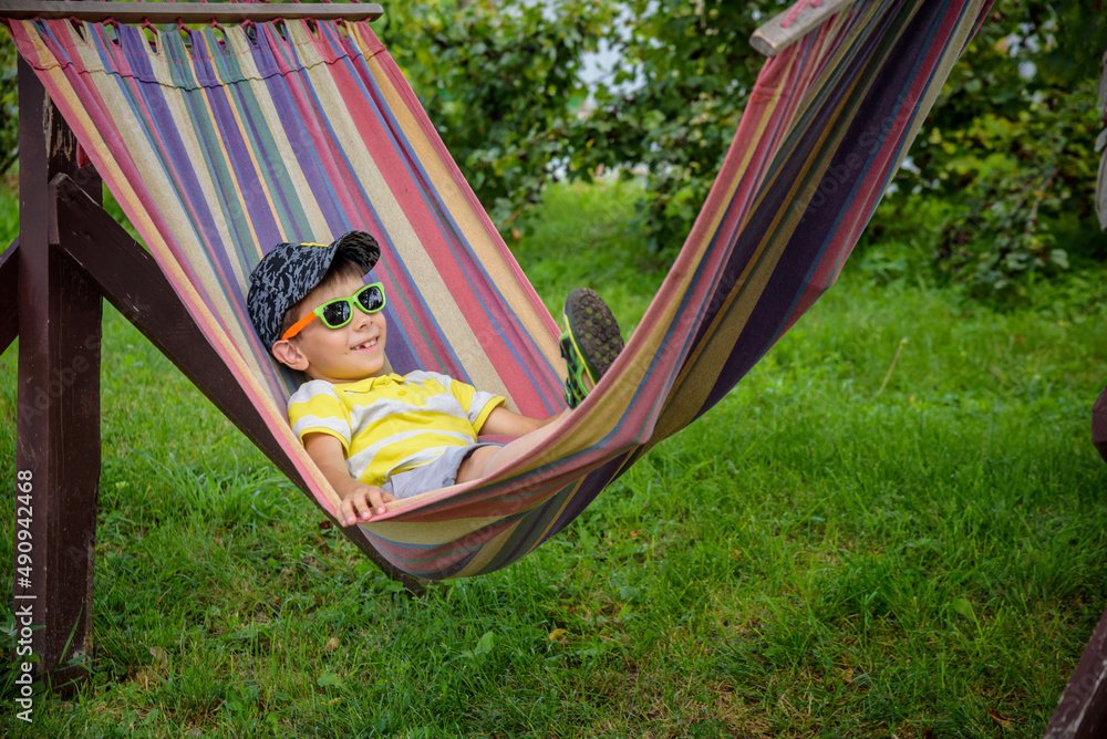 Cute little Caucasian boy relaxing and having fun in multicolored hammock in backyard or outdoor playground. Summer active leisure for kids. Child swinging on hammock. Activities for children Cute little Caucasian boy relaxing and having fun in multicolored hammock in backyard or outdoor playground. Summer active leisure for kids. Child swinging on hammock. Activities for children