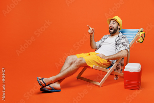 Canvas Print Young amazed tourist man in beach shirt hat lie on deckchair near fridge point finger aside on workspace isolated on plain orange background studio portrait