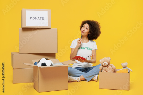 Full size young woman of African American ethnicity in white volunteer t-shirt hold boxes with presents write down list isolated on plain yellow background Voluntary free work assistance help concept photo