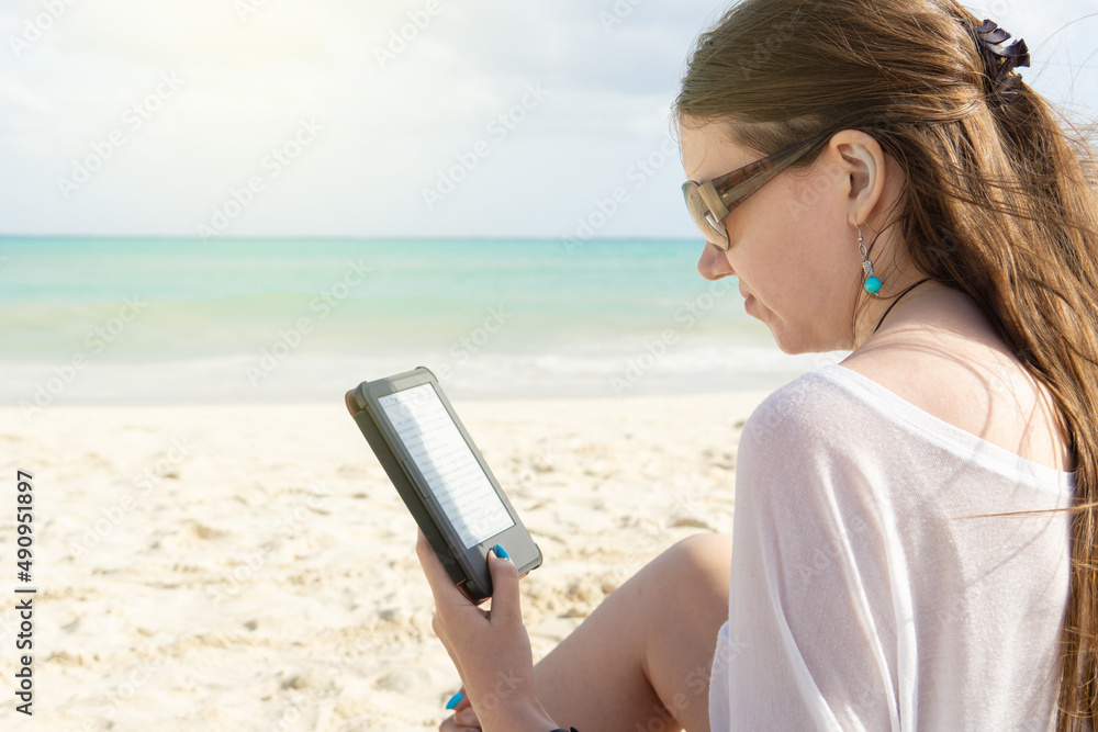 A girl in sunglasses reads an e-book on a sandy beach. Girl with an e-reader on the seashore.