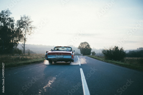 A retro car drives along the road at dawn. The convertible drives off into the distance on a deserted road