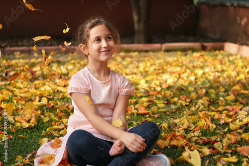 A little girl with Smiley face is sitting on yellow autumn leaves, leaves are falling on top of her.