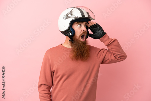 Young reddish caucasian man with a motorcycle helmet isolated on pink background doing surprise gesture while looking to the side