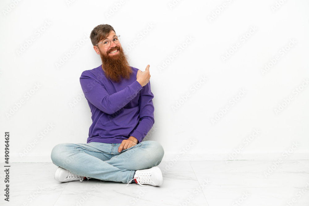 Young caucasian reddish man sitting on the floor isolated on white background pointing back Young caucasian reddish man sitting on the floor isolated on white background pointing back