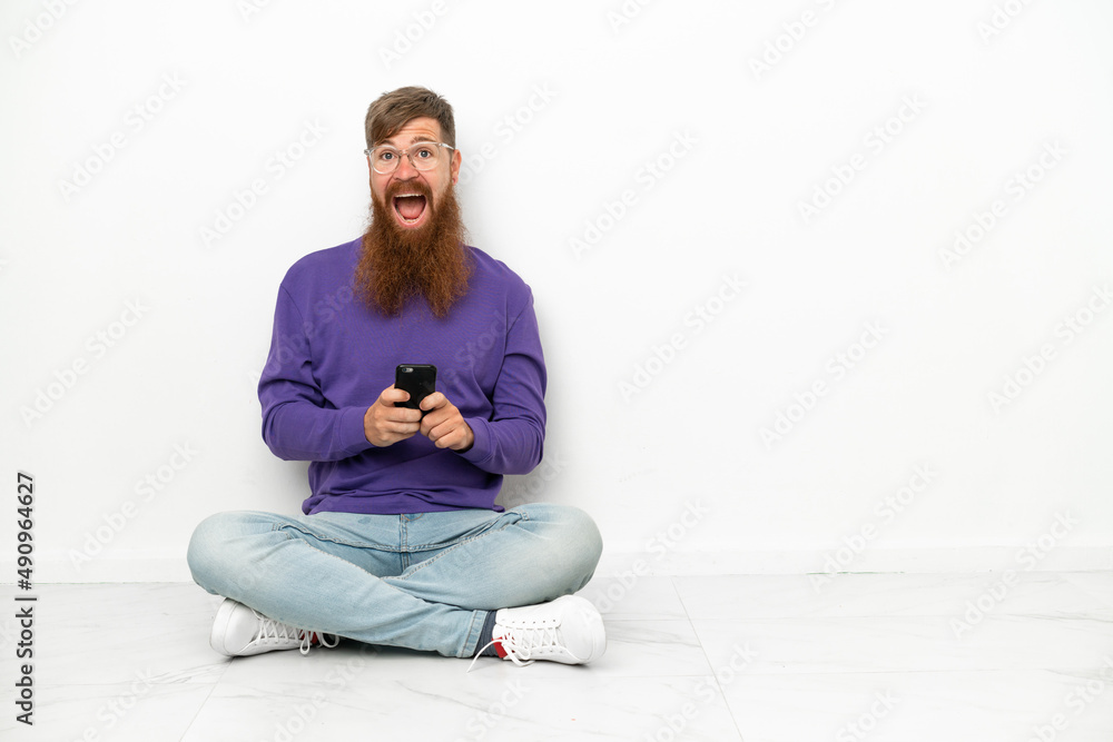 Fototapeta premium Young caucasian reddish man sitting on the floor isolated on white background surprised and sending a message