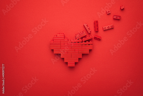Red falling apart heart symbol made of plastic building blocks. Flat lay image of breaking down like button on red background. Minimalist photo of stylized dissolving love symbol. photo