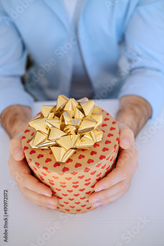 Man's hand holding a gift box with hearts present with a gold bow. Delivery Express, Sale. Online shopping. St. Valentine's Day International Women's Day birthday holiday concept. Vertical photo 
