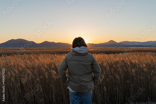 A woman standing in a reed field puts her hands in her pockets and looks at the sunset © ToriNim