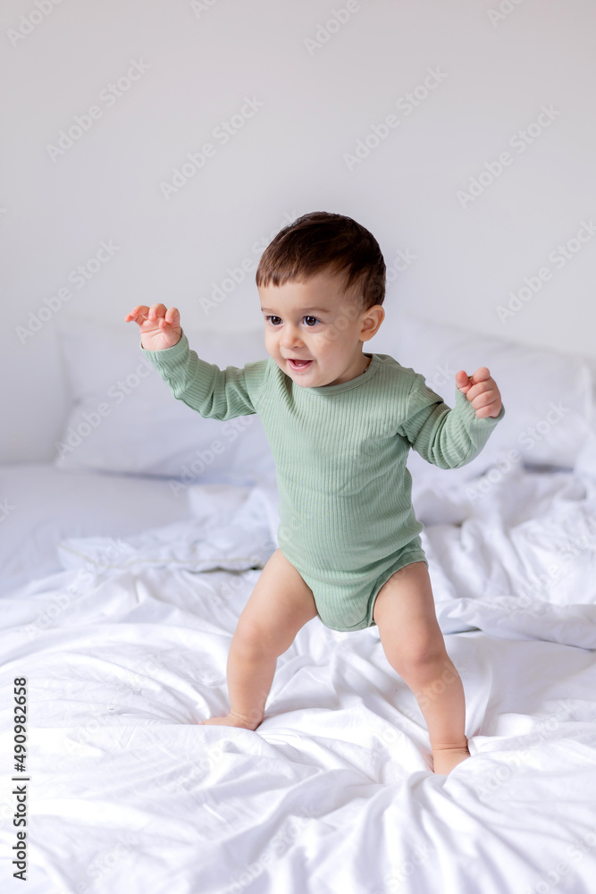 baby in a green cotton bodysuit stands barefoot on a white bed baby in a green cotton bodysuit stands barefoot on a white bed