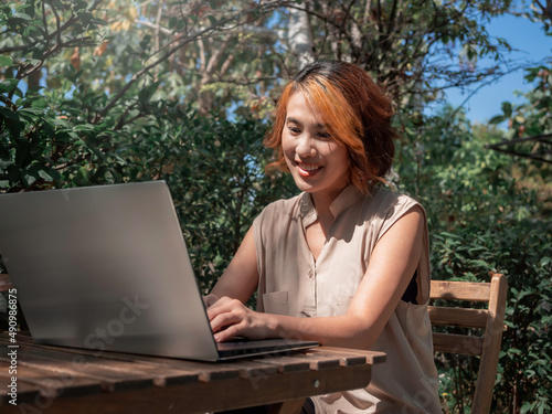 фотографія Attractive happy Asian woman portrait with trendy short hair working, looking at a silver laptop computer screen on wooden desk with a relaxing smile in the outdoor garden