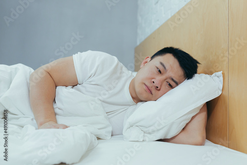 Sad Asian lying in a bed under a blanket, a depressed man in the morning