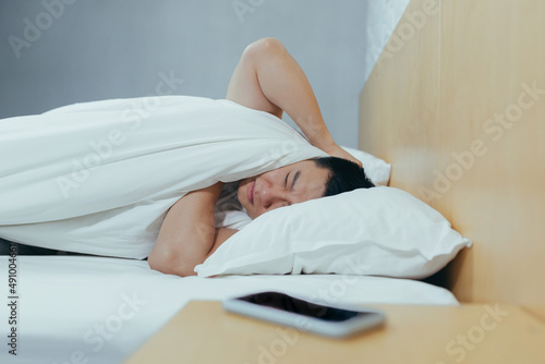 A man at home tries to fall asleep, noisy neighbors interfere with sleep, an Asian closes his ears with pillows, tired after work