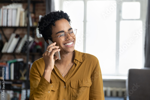 Cheerful young Black entrepreneur girl in stylish glasses talking on mobile phone with happy toothy smile, speaking on cellphone to client, negotiating on business project, enjoying telephone call