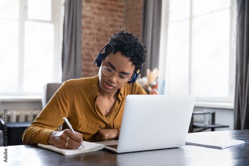 Focused Black student girl in glasses and headphones writing notes at laptop, checking handwriting summary, watching webinar, listening learning audio lecture, taking online virtual training course Focused Black student girl in glasses and headphones writing notes at laptop, checking handwriting summary, watching webinar, listening learning audio lecture, taking online virtual training course
