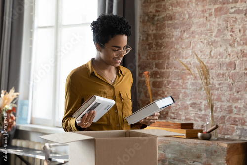Positive happy African student girl unpacking box with books, receiving parcel with purchase from bookstore, opening cardboard package, paper container, moving, relocating in new home