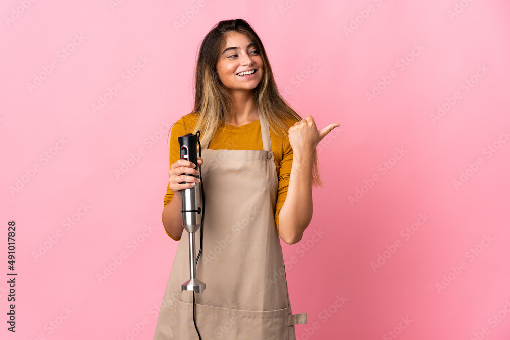 Young chef woman using hand blender isolated on pink background pointing to the side to present a product Young chef woman using hand blender isolated on pink background pointing to the side to present a product