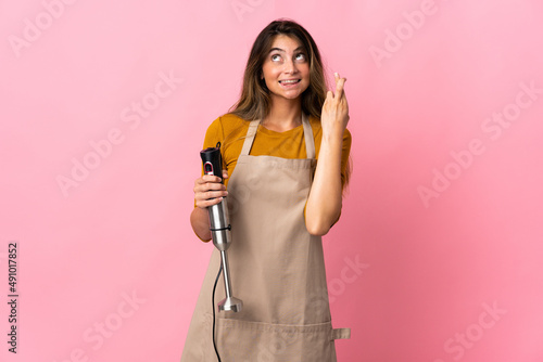 Young chef woman using hand blender isolated on pink background with fingers crossing and wishing the best