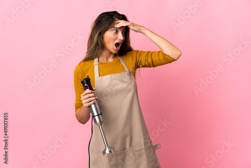 Young chef woman using hand blender isolated on pink background doing surprise gesture while looking to the side