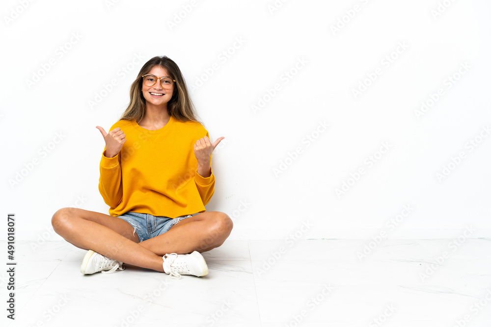 Young woman sitting on the floor isolated on white background with thumbs up gesture and smiling Young woman sitting on the floor isolated on white background with thumbs up gesture and smiling