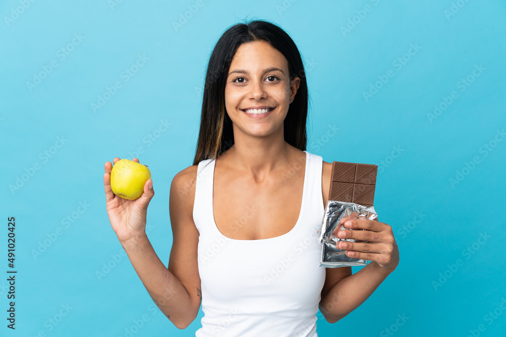 Caucasian girl isolated on blue background taking a chocolate tablet in one hand and an apple in the other Caucasian girl isolated on blue background taking a chocolate tablet in one hand and an apple in the other