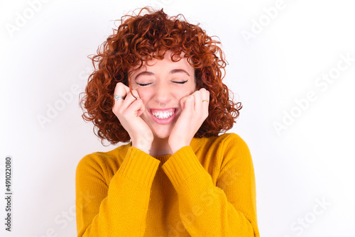 Portrait of young redhead girl wearing yellow sweater over white background being overwhelmed  expressing excitement and happiness with closed eyes and hands near face.