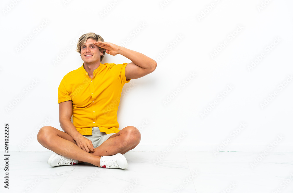 Fototapeta premium Young English man sitting on the floor saluting with hand with happy expression