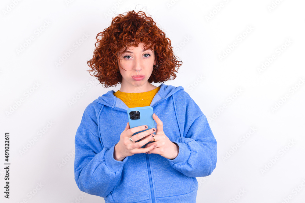Portrait of a confused young redhead girl wearing blue jacket over white background holding mobile phone and shrugging shoulders and frowning face. Portrait of a confused young redhead girl wearing blue jacket over white background holding mobile phone and shrugging shoulders and frowning face.