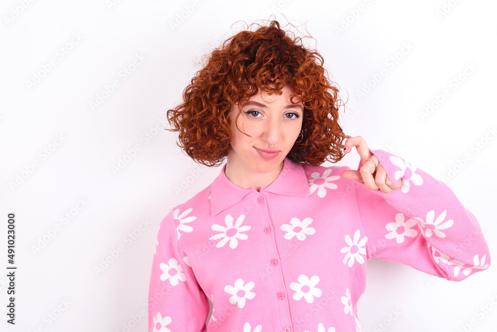 Displeased young redhead girl wearing pink floral t-shirt over white background shapes little hand sign demonstrates something not very big. Body language concept. Displeased young redhead girl wearing pink floral t-shirt over white background shapes little hand sign demonstrates something not very big. Body language concept.