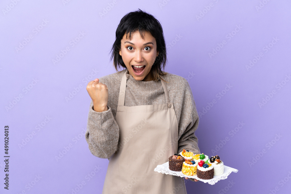 Young Uruguayan woman holding lots of different mini cakes over isolated purple background celebrating a victory