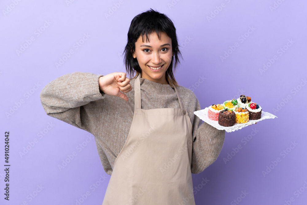 Young Uruguayan woman holding lots of different mini cakes over isolated purple background proud and self-satisfied Young Uruguayan woman holding lots of different mini cakes over isolated purple background proud and self-satisfied