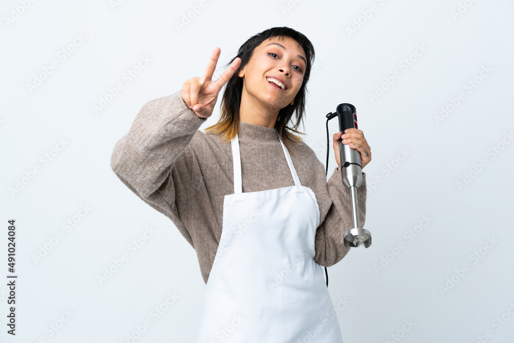 Chef Uruguayan girl using hand blender over isolated white background smiling and showing victory sign Chef Uruguayan girl using hand blender over isolated white background smiling and showing victory sign