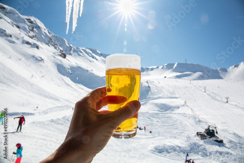 A person is holding a glass of beer with white foam in the mountains with snow on a sunny day. A tourist with a beer in a ski resort