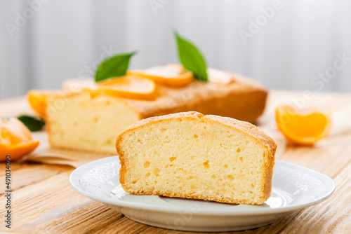 Close up piece of moist orange fruit pound cake on plate with slices of orange and whole pie on background. Delicious breakfast, traditional English tea time. Recipe of orange pie loaf.