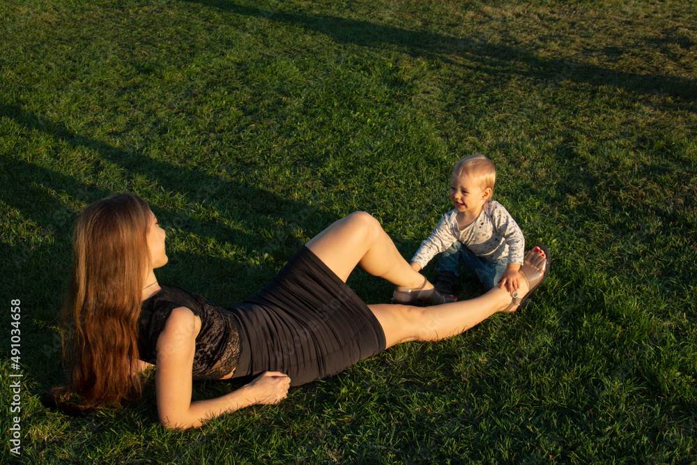 Little toddler son playing with mother legs, woman with her child outdoor on the grass lawn Little toddler son playing with mother legs, woman with her child outdoor on the grass lawn