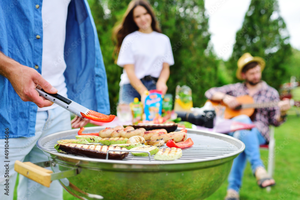 man's hand holds a barbecue tongs with a juicy delicious meat steak against the background of a barbecue grill with meat and vegetables and a group of friends on a picnic who are having fun and man's hand holds a barbecue tongs with a juicy delicious meat steak against the background of a barbecue grill with meat and vegetables and a group of friends on a picnic who are having fun and